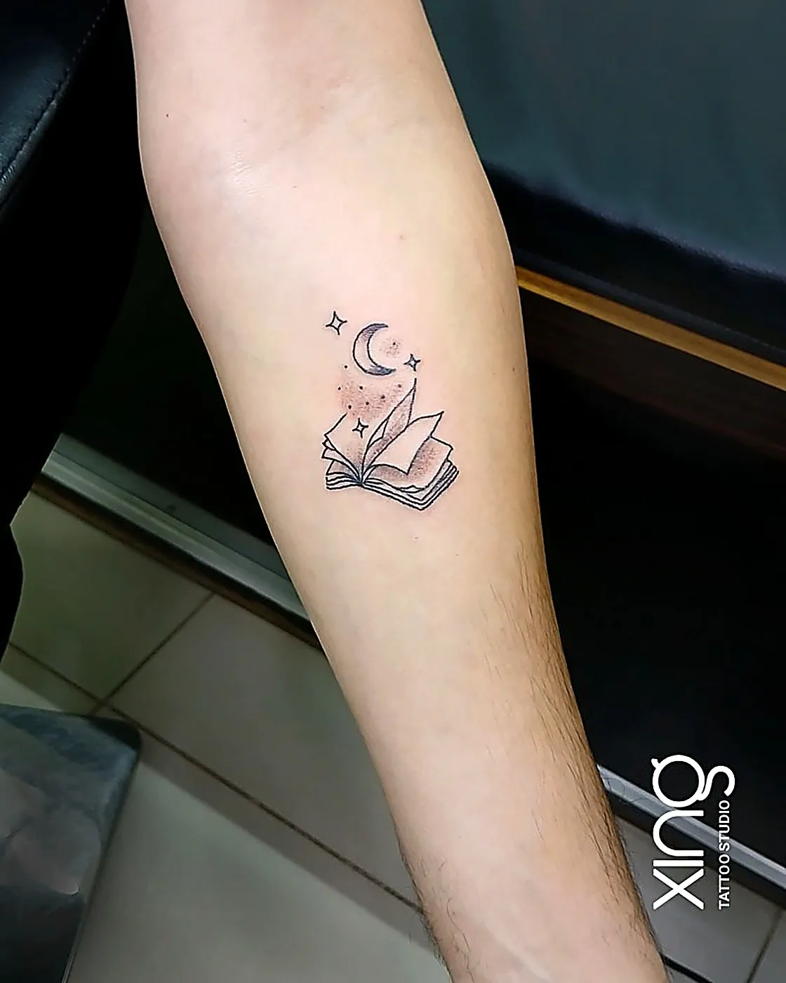 Fine-line open book with moon and stars tattoo on forearm